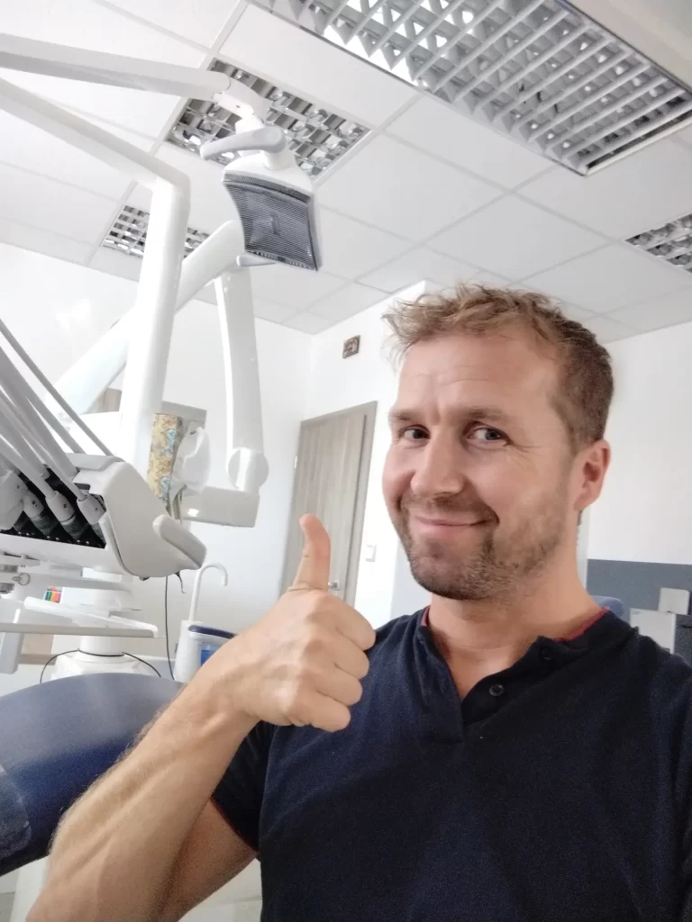 Muž dává palec nahoru v zubní ordinaci v Uherském Hradišti, usmívá se s dentálním vybavením viditelným v pozadí.